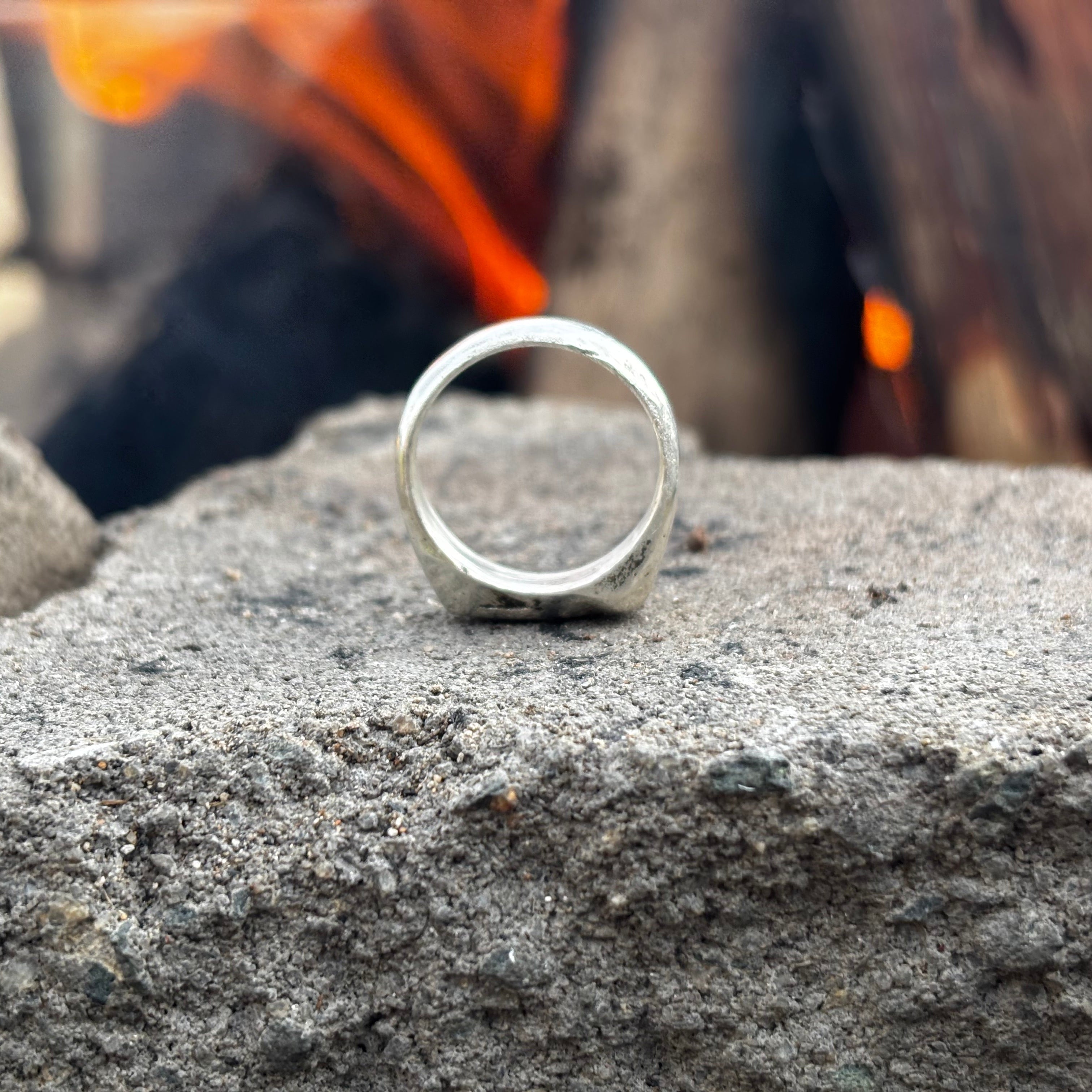 A silver band ring with a textured, hammered finish resting on a rough gray stone, with a blurred background of fire and dark wood.